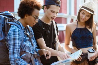 Group of Young Travelers Exploring with a Map Three young travelers are examining a map. One has a backpack and plaid shirt, another wears a black cap, and the third has a hat and holds a coffee cup.
