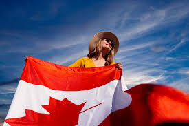 Woman Proudly Holding Canadian Flag Outdoors A woman is standing outdoors, holding a large Canadian flag. She is wearing a hat and sunglasses, looking upward, with a clear blue sky in the background.